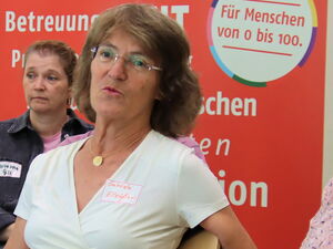 4 Frauen in einem Raum, sie sitzen versetzt auf Stühlen. Eine ältere Frau mit Notizen auf dem Schoß spricht. Die anderen sehen interessiert zu ihr. Die Frau im Zentrum trägt Brille, eine goldene Kette und ein weißes T-Shirt mit tiefen V-Ausschnitt.