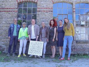 Gruppenbild: 7 Menschen vor einem alten Gebäude mit vielen kleinen Fensterscheiben. Ein Mann und eine Frau in der Mitte halten ein Bild.