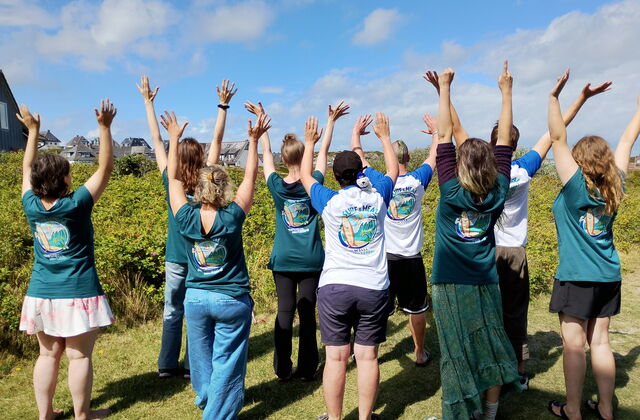 Gruppenbild, 8 Personen von hinten. Alle tragen ein "Surf and Heal" T-Shirt und strecken die Arme nach oben. Sie stehen auf einer Wiese.