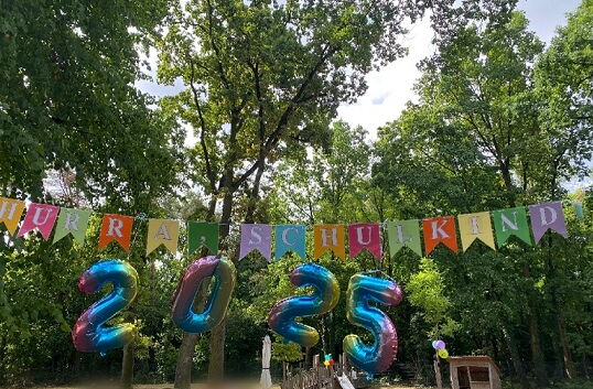 Luftballons in Form der Zahl „2025“ und bunte Wimpelgirlande hängen an Bäumen im Freien.