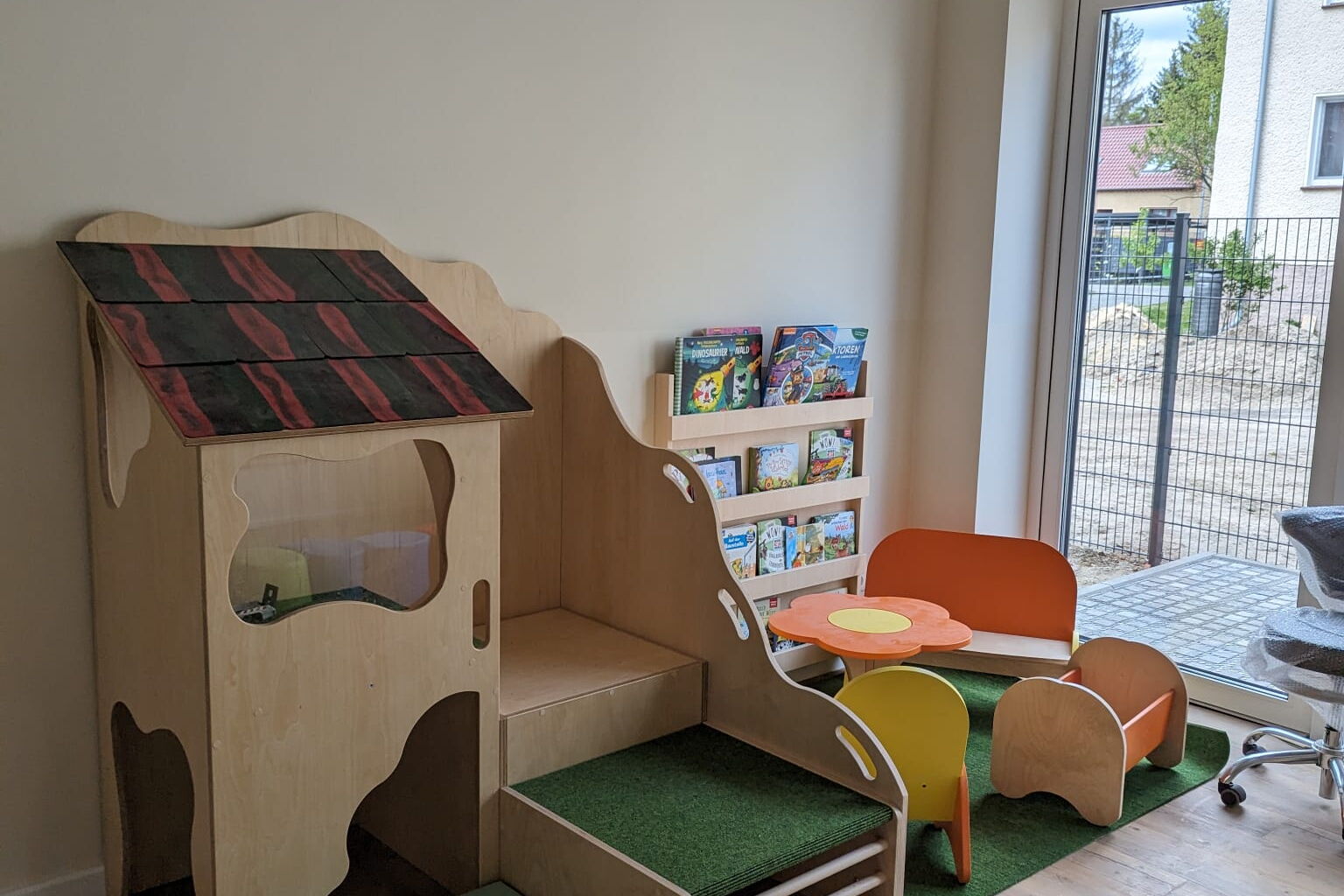 Raum. Bodentiefes Fenster, Spielbereich. Mit kleiner Puppen-Sitzecke und Puppenbett. Und ei kleines Kita Kletterhaus.