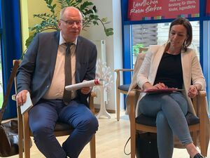 Ein Raum. Ein Mann und eine Frau sitzen auf Stühlen. Der Mann ist schon älter, hohe Stirn und graue Haare mit Brille, weißem Hemd, grauen Schlips und grauer Anzugjacke, die Frau jünger, dunkle glatte Haare macht sich Notizen.