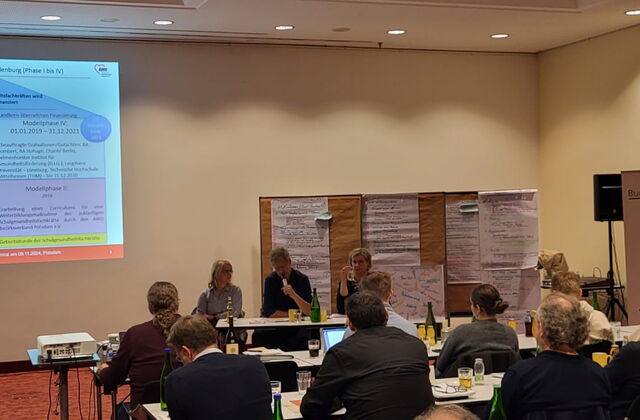 Konferenzraum. Teilnehmer sitzen an Tischen mit Getränken. Eine Präsentation wird an die Wand geworfen. 3 Personen sitzen den Teilnehmern zugewandt, die Person in der Mitte spricht in ein Mikrofon.
