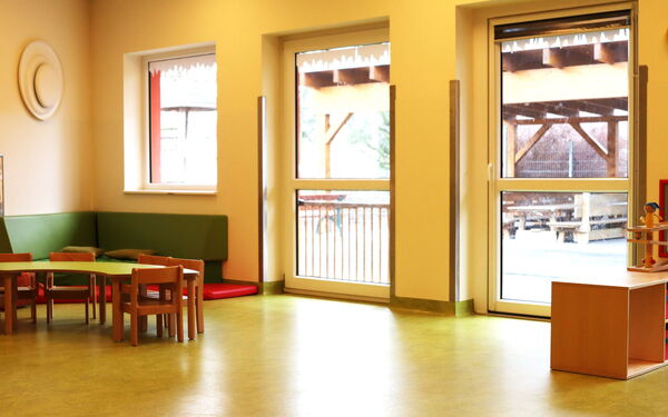 Heller Raum, kleine Tische und Stühle in der Ecke eine grüne Sitzecke. Rechts im Bild kleine Regale mit Spielzeug. Große Fenster lassen Licht hinein.