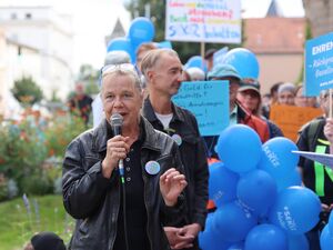 Blonde Frau hat Brille auf den Kopf geschoben und spricht in ein Mikrofon. Sie trägt eine schwarze Lederjacke mit Button der Aktion. Um sie rum Menschen mit blauen SEKIZ Luftballons und Protestschildern.