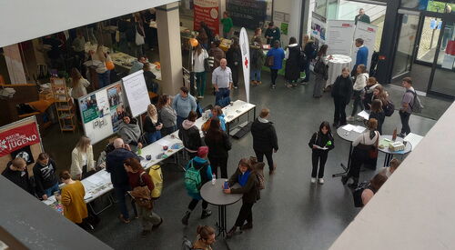 Ein Foyer von schrägoben. Stehtische und Info-Stände. Mehrere Menschen stehen an den Tischen und stehen im Raum herum.