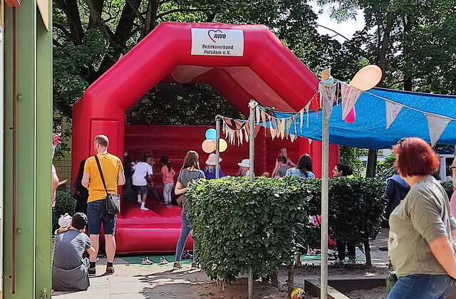 Rote Hüpfburg oben mit AWO Potsdam Logo. Kinder sind auf der Hüpfburg. Kinderschuhe stehen davor. Es ist ein sonniger Tag.