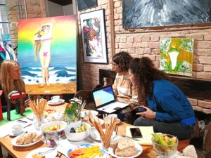 3 Frauen sitzen in einer Galerie. Vor ihnen ein reichgedeckter Tisch, an den Wänden Bildern. In der Ecke ein großes Bild auf eine Staffelei. Ein Bild an der Wand zeigt den Froschkönig. Frosch mit Krone.