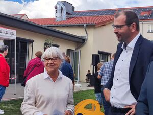 Draußen, ältere Frau mit weißen Haaren und zwei Männern in Anzug. Im Hintergrund Erwachsene und Kinde, die das Gelände erkunden.