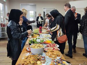 Drinnen. Langer Tisch mit üppigem Büfett, Salate, Kuchen, Muffins, Gemüse- und Obstteller. Menschen stehen auf beiden Seiten des Tisches und bedienen sich.