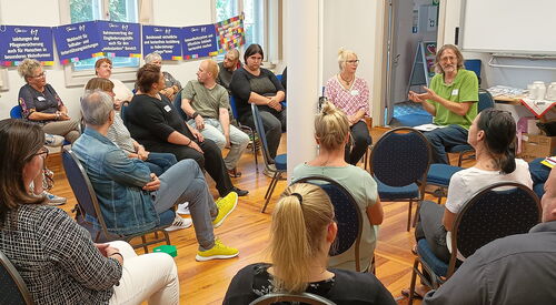 Großer Raum. Die Stühle sind in mehreren Kreisen angeordnet. Im inneren Kreis wird lebhaft diskutiert. An der Fensterwand hängen Schilder mit Forderungen der AWO.