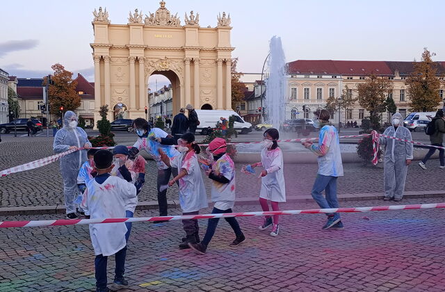 Der Luisenplatz mit Springbrunnen und brandenburger Tor im Hintergrund. Ein Absperrband wird von Menschen in Schutzanzügen / Masken gehalten sodass ein Rechteck umspannt ist, darin Mädchen in weißen Shirts bunt befleckt, auch der Steine / sind kunterbunt.