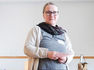 Frau steht in einem Raum und schaut in die Kamera, sie trägt Brille, dunkles Halstuch, helle Strickjacke über einen grauen Oberteil. Sie hält ein Blatt Papier in den Händen.