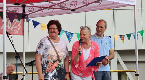 2 Frauen und ein Mann auf der Bühne, alle gut gelaunt. Frau in der Mitte hat ein Mikrofon in der rechten Hand und in der linken ein Klemmbrett.