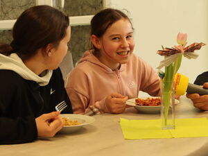 2 Mädchen am nebeneinander am Tisch beim Essen. Ein gelbes Antenne Mikrofon wird dem einen Mädchen hingehalten das in die Kamera lächelt.