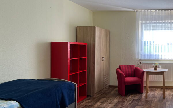 Bewohnerzimmer: vorn links ein Bett an der Wand weiter hinten ein rotes offenes Regal und ein Kleiderschrank in der Ecke des Zimmers. Ein roter Sessel und ein kleiner runder Tische stehen vor dem Fenster.