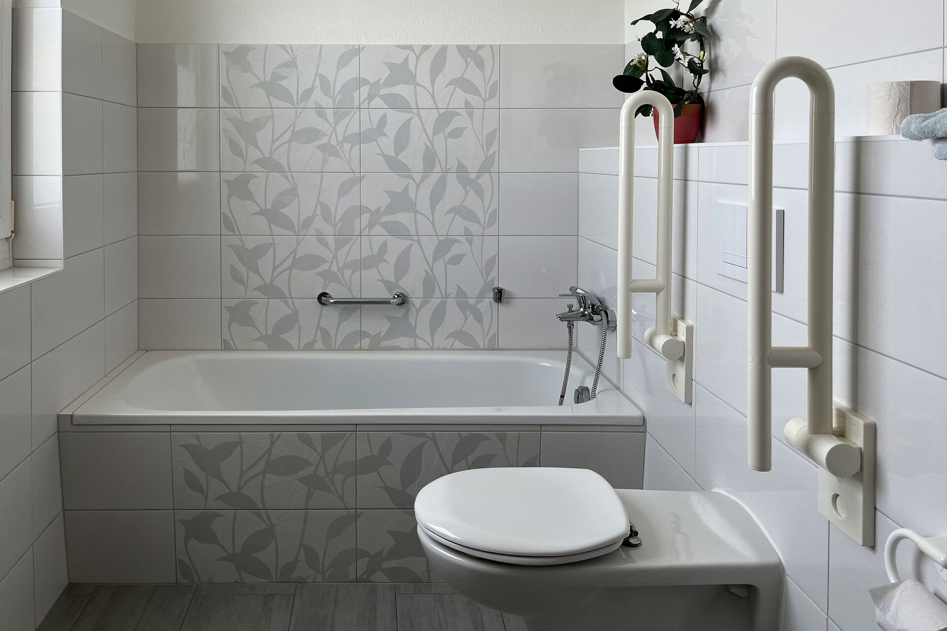 Badezimmer: Wanne mit Haltegriff an der Wand. WC mit hochgeklappten Haltegriffen rechts und links neben dem WC montiert.