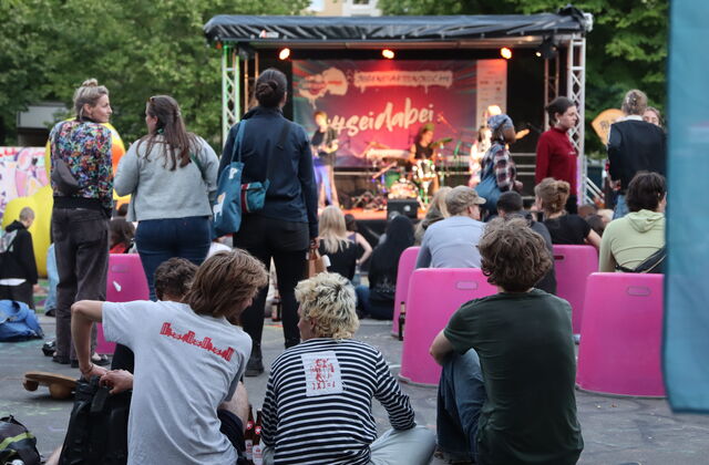 Junges Publikum sitzt auf dem Boden, in pinken Hockern und steht vor der Bühne. Auf der Bühne eine Band.