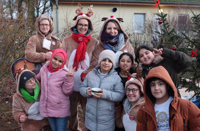 Vier weiße Frauen in Rentierkostümen. 3 stehen hinter südländischen Kindern in Winterkleidung. Eine der Frauen kniet zwischen den Kindern. Alle lächeln fröhlich in die Kamera. Eine Frau hat eine Gitarre umhängen.