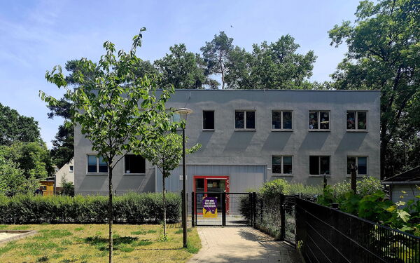 Zweistöckiges graues Gebäude. Mit einem verschlossenen Eingangstor. Vor den Gebäude: Wiese, Bäume und Hecke am Zaun.