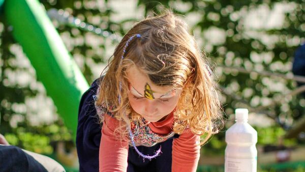 farbige Handabdrücke von Kindern auf einem Stofftuch. Ein Mädchen mit blonden Haaren drückt ihre flache Hand auf das Stofftuch, sie kniet auf dem Boden. Neben ihr ein Mann.