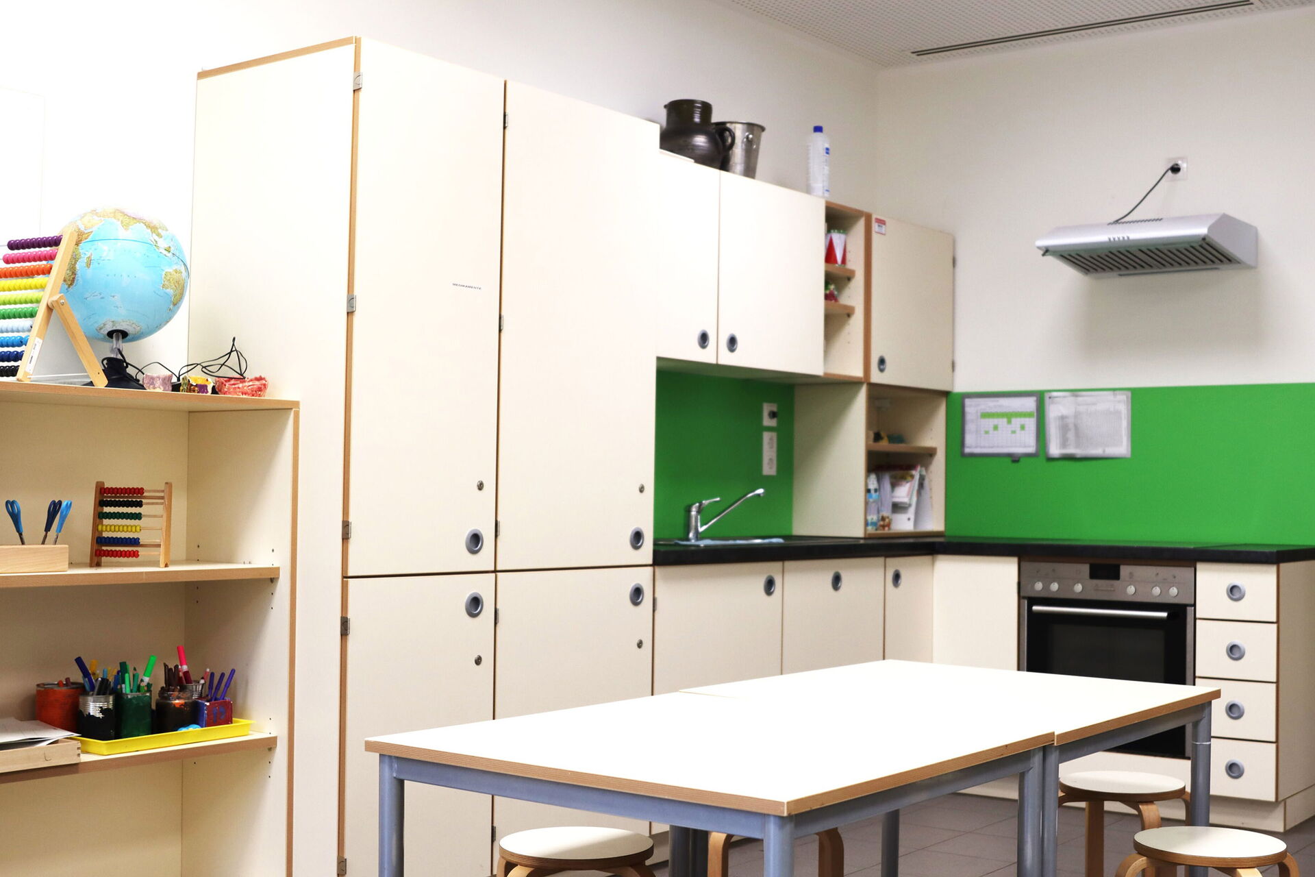 Eine Kinderküche mit großen Esstisch, Hockern, viel Stauraum durch Schränke, Herd, Abzugshaube und Spüle. Helle Möbel