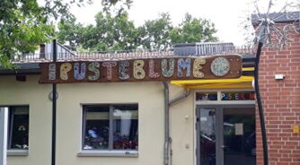 Flachbau mit Fenster und Eingangsbereich, Oben steht in farbigen Buchstaben Pusteblume am Gebäude.