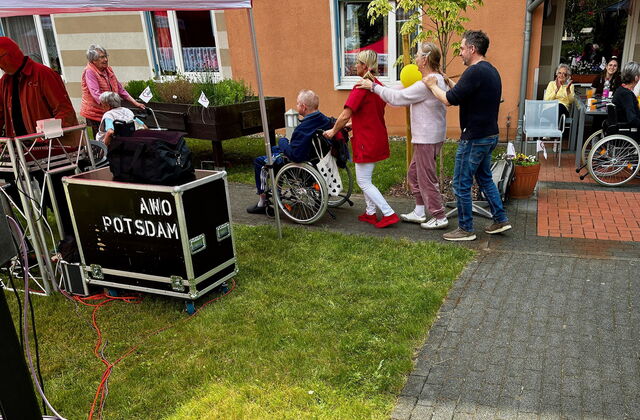 Polonäse: Draußen Sonne scheint. Vier Personen bilden eine Polonäse vor eine ältere Person im Rollstuhl.