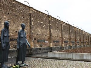 Draußen, eine Mauer oben mit Stacheldraht. Zwei Skulpturen stehen vor der Mauer. Die Skulpturen stellen Frauen in Kleidern da. Eine trägt eine Art Kapuze. Vor den grauen Skulpturen l