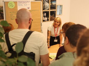 Teamarbeit. Flipchart an der Wand und eins auf eimem Tisch. Blonde Frau mit einem Stift, notiert Ideen der Teilnehmenden.