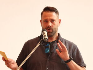Mann am Rednerpult. Er hat Karteikarten / Notizen in der rechten Hand und sprich in ein Mikrofon.