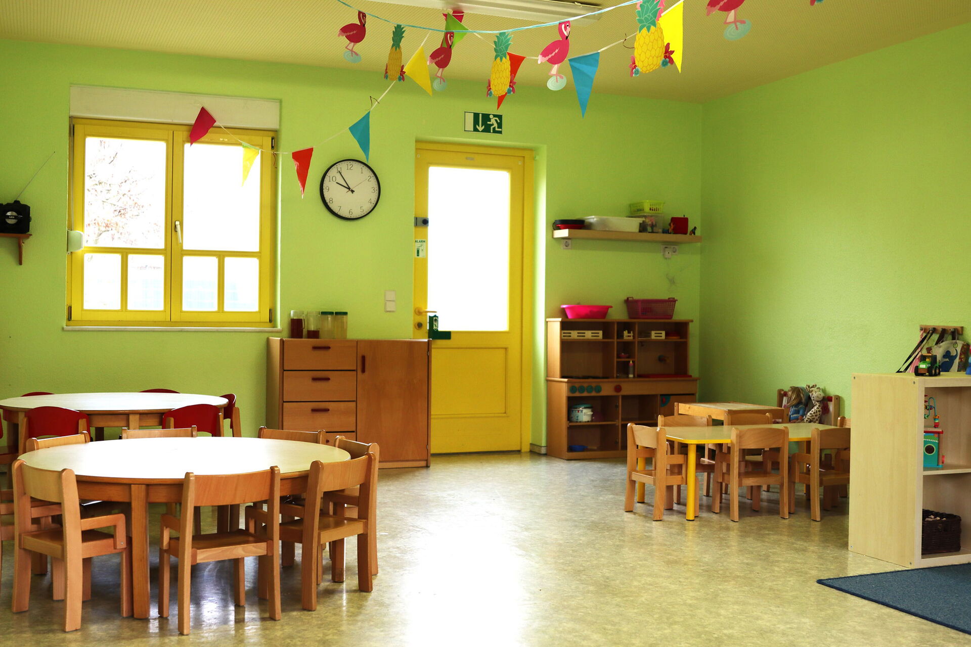 Spielraum. Zwei runde kleine Tische mit kleinen Stühlen, ein rechteckiger kleiner Tisch mit kleinen Stühlen, Regale, ein Schrank. An der Decke hängen Wimpel und eine Girlande mit Ananas und Flamingos.
