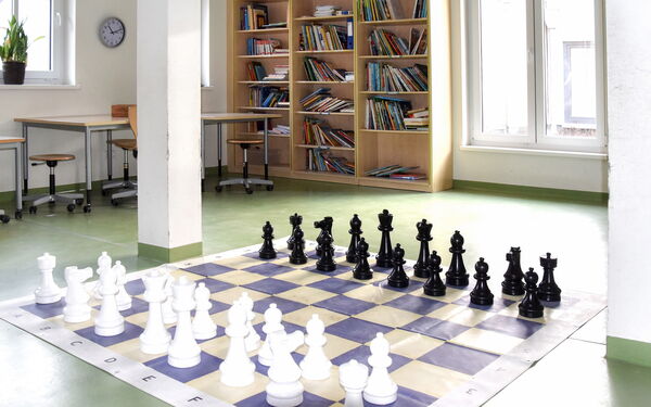 Ein Spielraum. Ein großes Schachspiel ist auf dem Fußboden aufgebaut. In der Zimmerecke ein Bücherregal mit Büchern.
