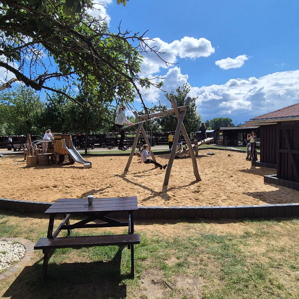 Spielplatz mit Schaukeln und Klettergerüst mit Rutsche für kleine Kinder. Davor Bänke mit Tisch.