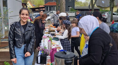 Draußen, ein Büfett, langer Tisch mit Getränken und Speisen. Kinder und Frauen holen sich was zu essen.