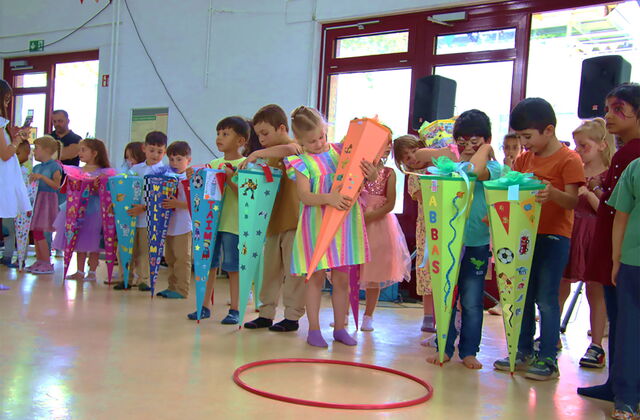 Ca. 12 Einschulungskinder mit ihren großen Zuckertüten in einem großen Raum. Die Zuckertüten sind farbig und bund beklebt. Die Kinder freuen sich.
