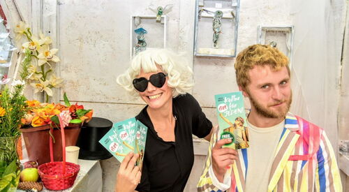 Zwei Menschen, Frau und Mann. Frau mit blonder wilder Frisur und Herz-Sonnenbrille hält drei Flyer von Kultur für Jeden in der Hand. Mann dunkelblond mit Bart einen Flyer von Kultur für Jeden in der Hand.