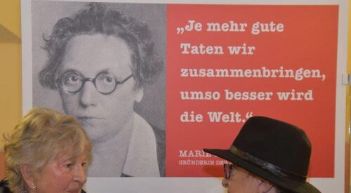 Hintergrund: Plakat mit schwarz-weiß Foto von Marie Juchacz mit Zitat: Je mehr gute Taten wir zusammenbringen, umso besser wird die Welt. Davor zwei ältere Frauen im Gespräch.
