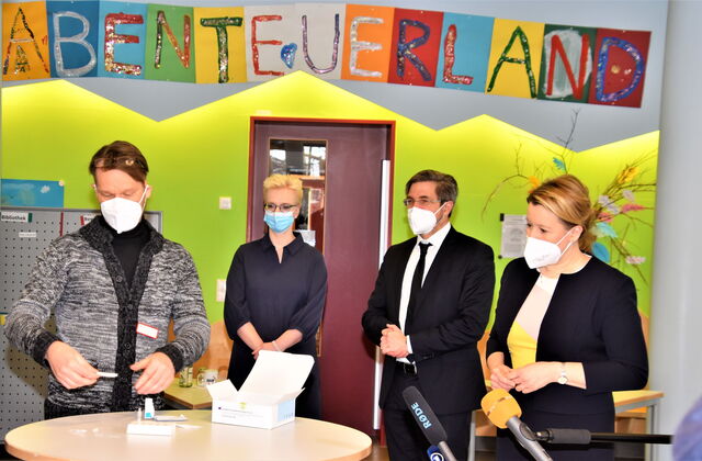 Ein farbenfroher Raum, grüne Wand. Vier Personen mit FFP 2 und OP Masken stehen um einen runden Stehtisch. Darauf eine Packung mit Corona Tests. Darüber bunte Buchstaben auf farbigen Papier: ABENTEUERLAND