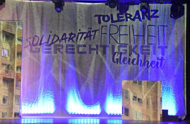 Bühnenvorhang mit Text: Toleranz, Solidarität, Freiheit, Gerechtigkeit, Gleichheit. Dahinter wird die Kulisse des Theaterstücks sichtbar. Ein Häuserblock.