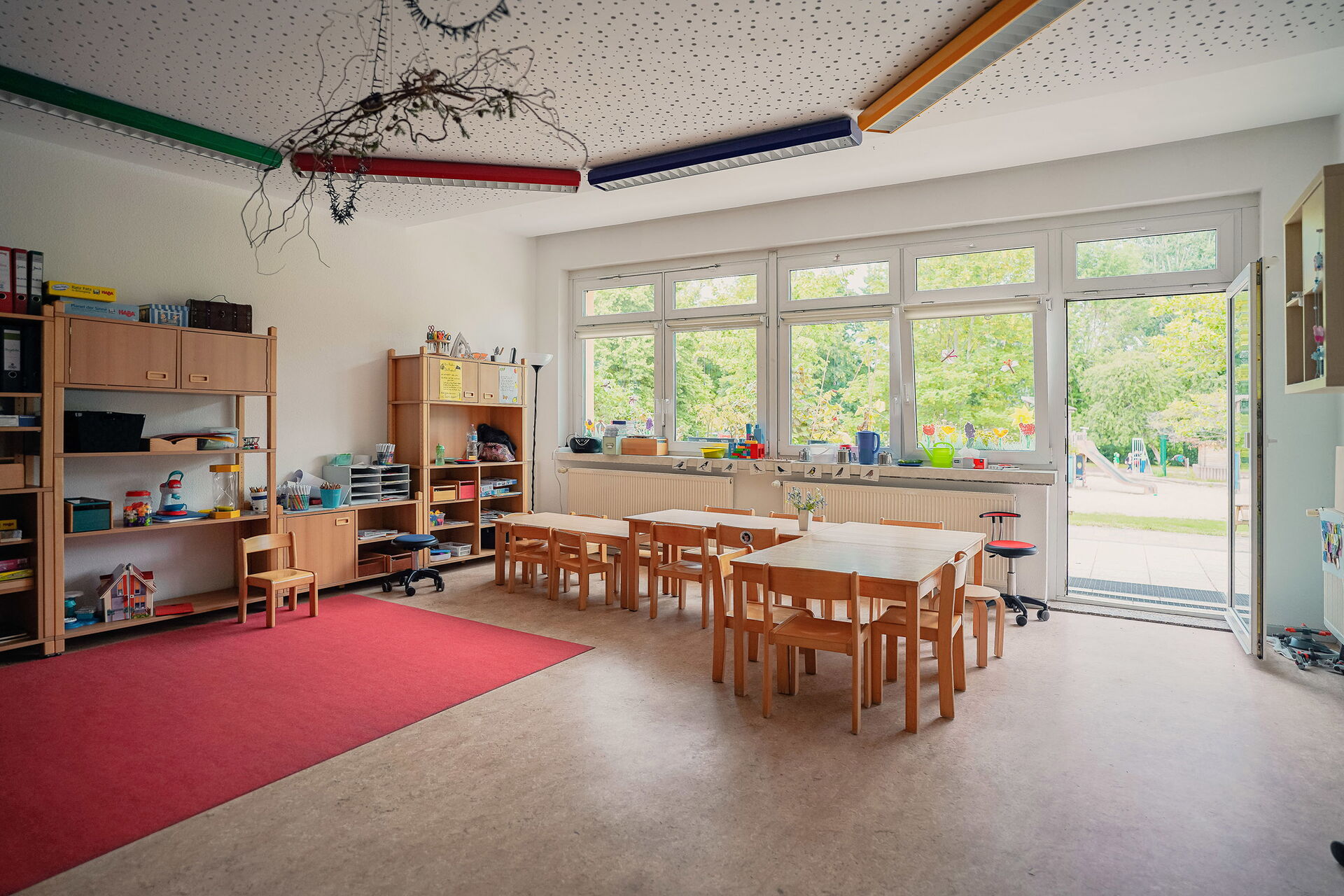 Großer heller Raum, die Terrassentür steht offen. Im Raum Tische mit Stühle, Regale mit Spielzeug an der Wand.