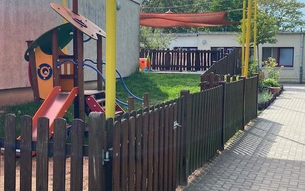 Gebäude. Draußen, ein kleines Klettergerüst mit Treppe uns Rutsche für Kleinkinder. Darüber ist ein organgenes Sonnensegel gespannt. Ein Holzzaun geht um das Kita-Gelände.