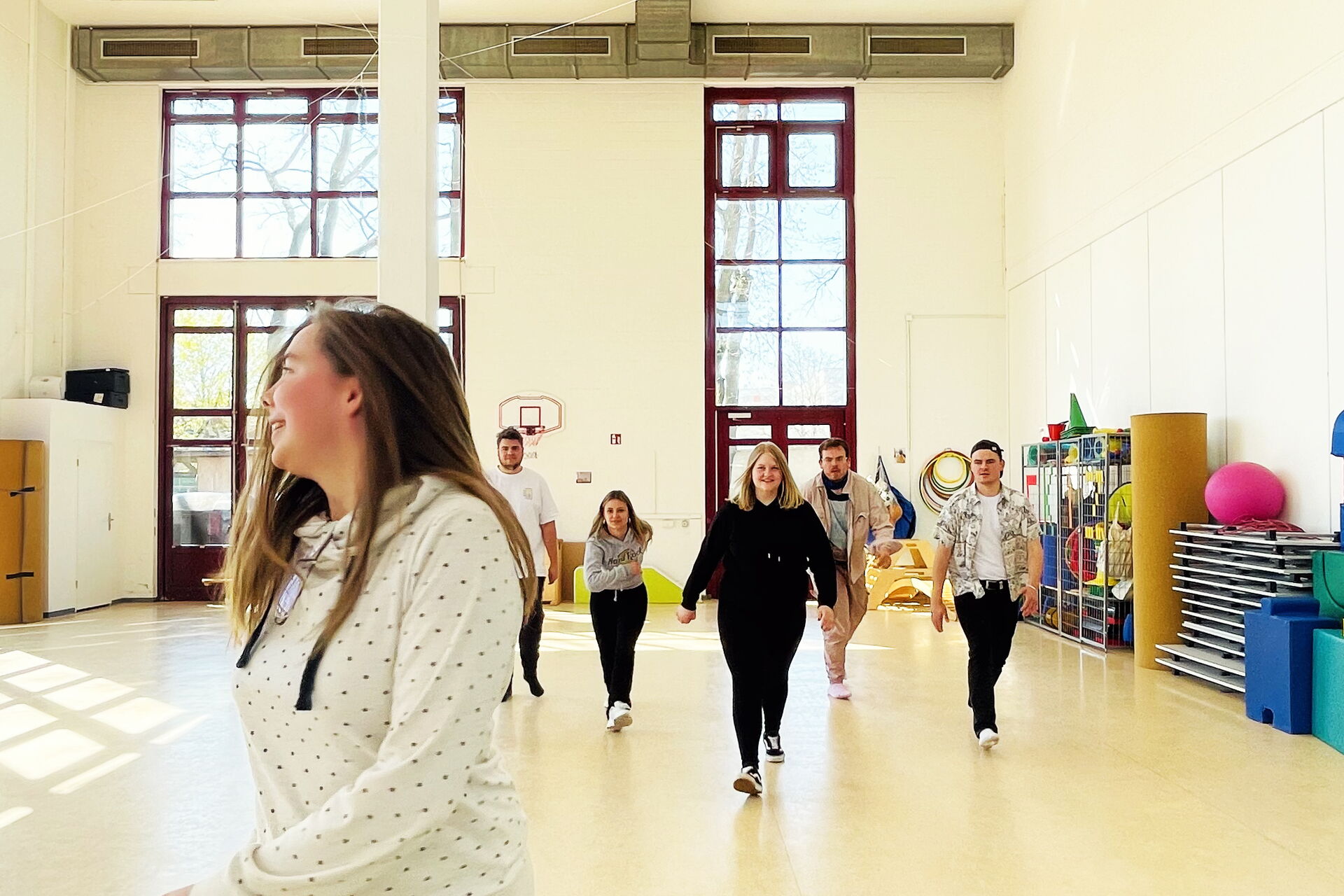 Eine Turnhalle  mit hellen Wänden. Junge Menschen schreiten durch die Halle. Kommen auf die Kamera zu.