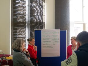 Ein Raum mitt großen Fenstern, mit grauen Vorhängen abgedunkelt. Zwei Frauen stehen vor einem Flipchart das mit grüner Schrift beschrieben ist. Wenige weitere Person stehen davor.