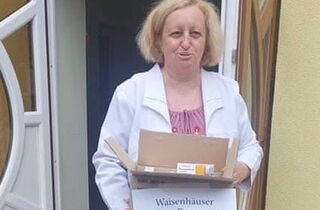 Auf dem Bild ist eine Frau mit weißem Kittel, vor einem ukrainischen Waisenhaus. Sie hält eine Kiste mit gespendeten Medikamenten in der Hand