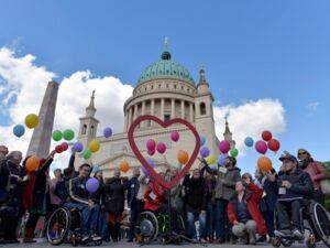 Vor der Nikolaikirche Potsdam, VViele Menschen mit und ohne Behinderung, u.a. drei Rollstuhlfahrer. In der Mitte ist ein großes Herz aufgestellt, und die Menschen lassen bunte Luftballons fliegen.