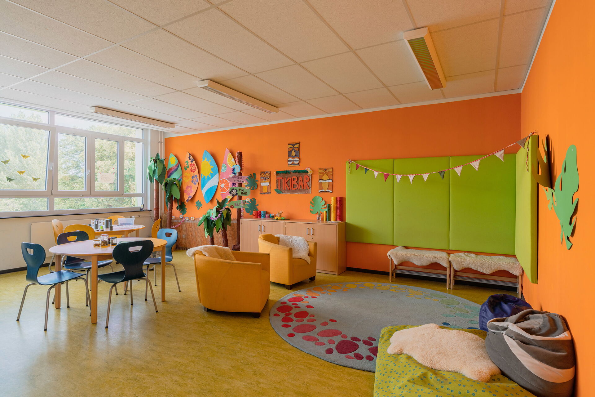 Raum mit orangene Wänden, lichtspendenen Fenster, im Raum ein großer runder Teppisch, zwei gelbe Sessel, zwei Tische mit je 4 Stühlen vor den Fenstern. Gebastelte Wanddekoration.