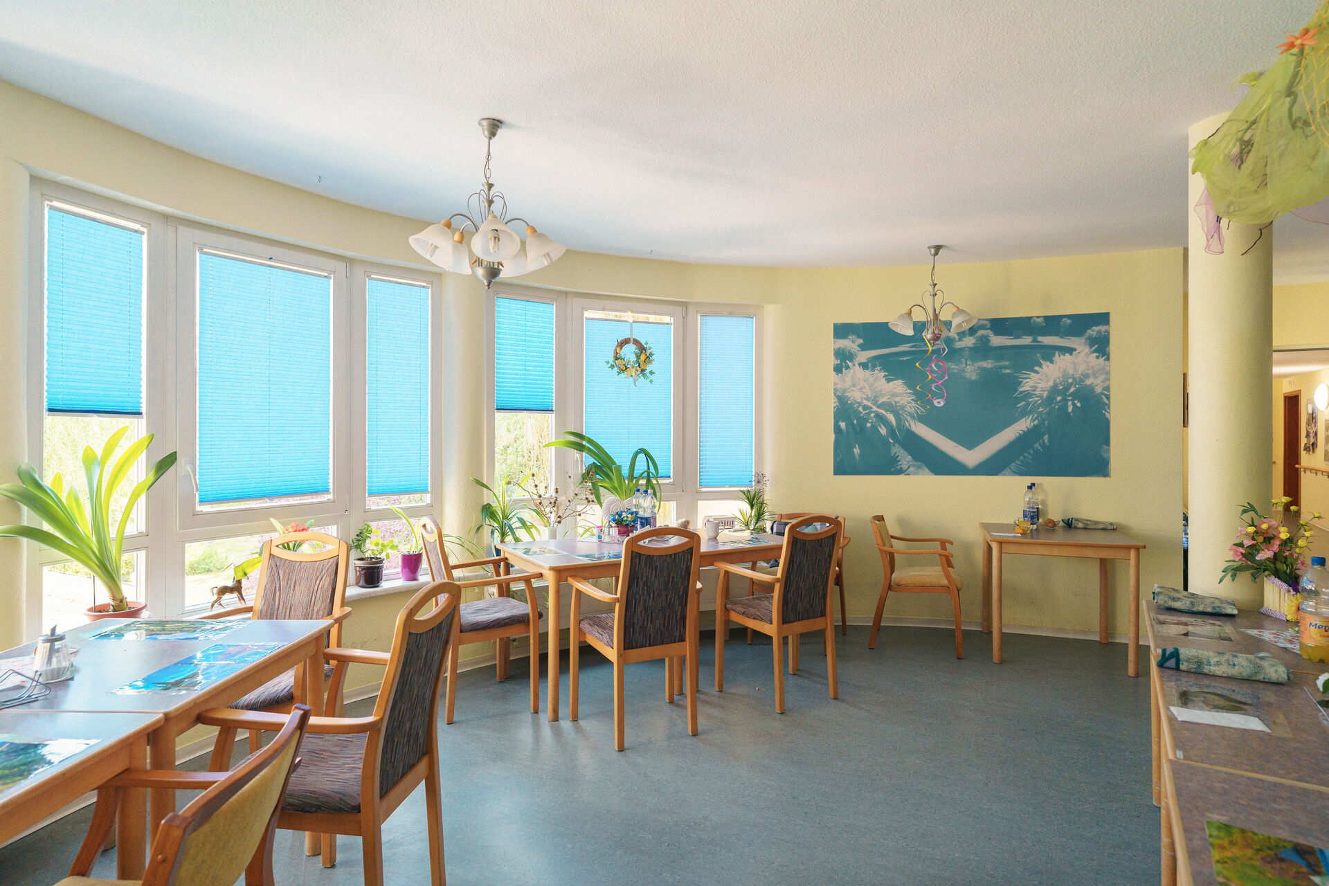 kleiner Speisesaal. Freundliche hellgelbe Wände, Tisch mit Stühlen stehen außen wo viele Fenster sind.