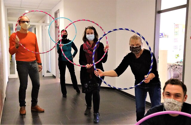 Vier Personen in einem Flur. Alle tragen Stoffmasken. Alle halten bunte Hula Hoop Reifen hoch.