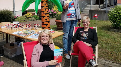 Ein Haus, im Vordergrund zwei Frauen sitzen in roten Sonnenstühlen unter einer aufblasbaren Gummipalme. Eine Frau steht hinter ihnen mit einem Fächer in der Hand.